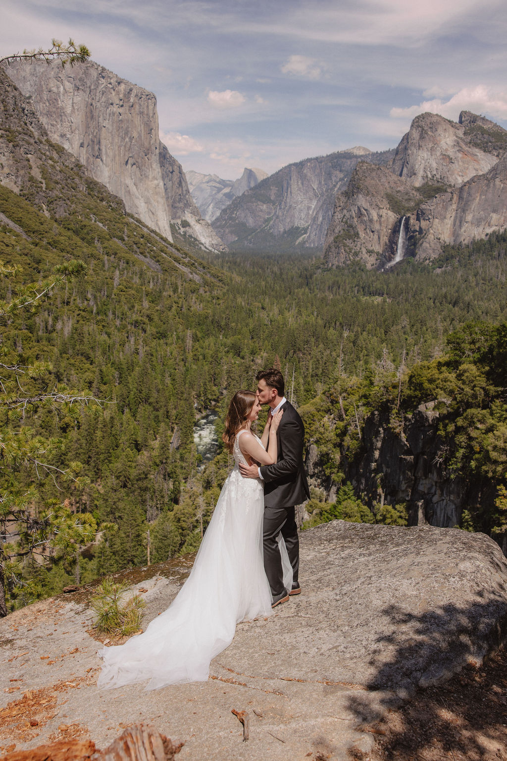 A bride and groom in wedding attire stand on a rocky overlook, embracing while facing a scenic view of mountains, cliffs, and forest in Yosemite National Park.