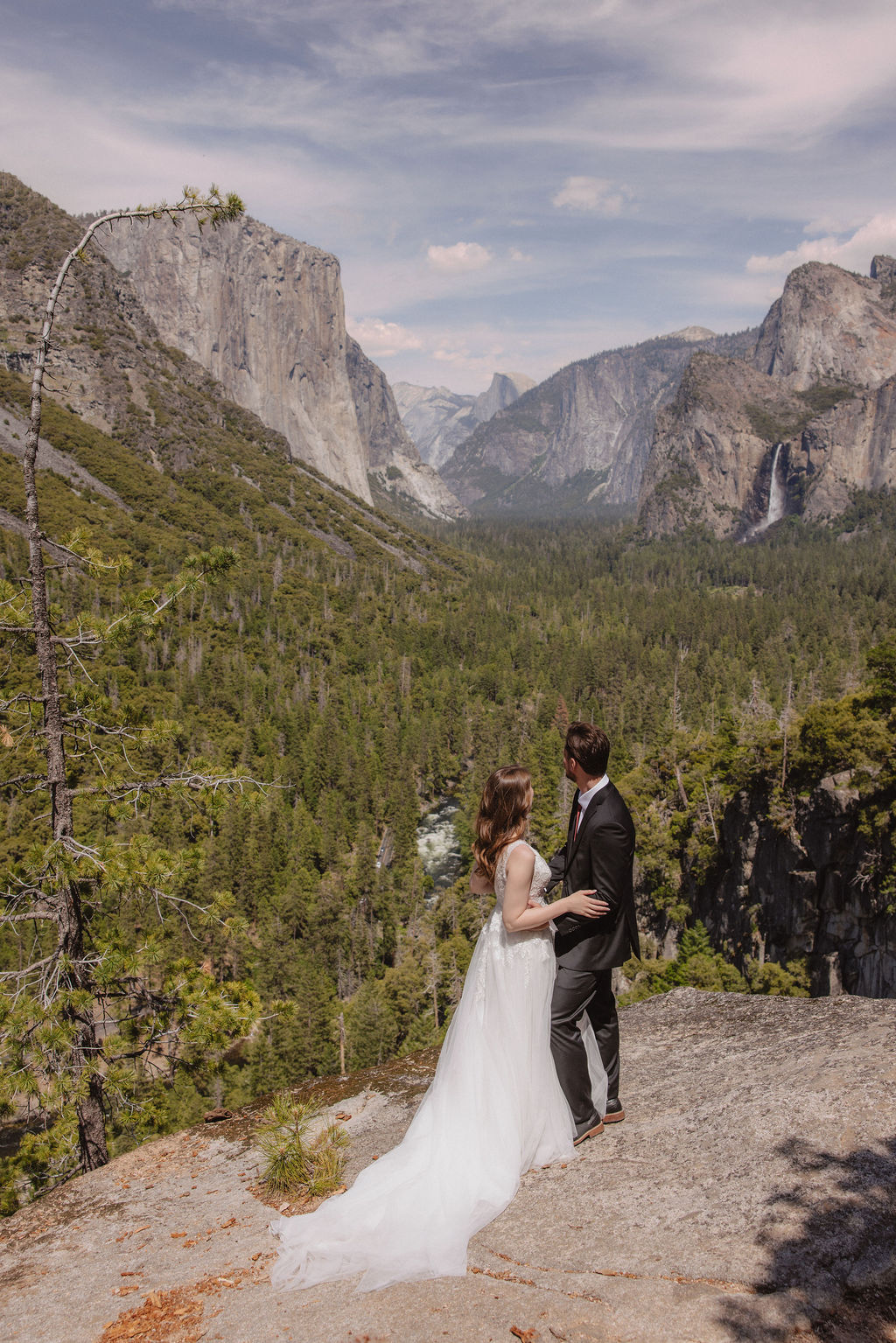 A bride and groom in wedding attire stand on a rocky overlook, embracing while facing a scenic view of mountains, cliffs, and forest in Yosemite National Park.