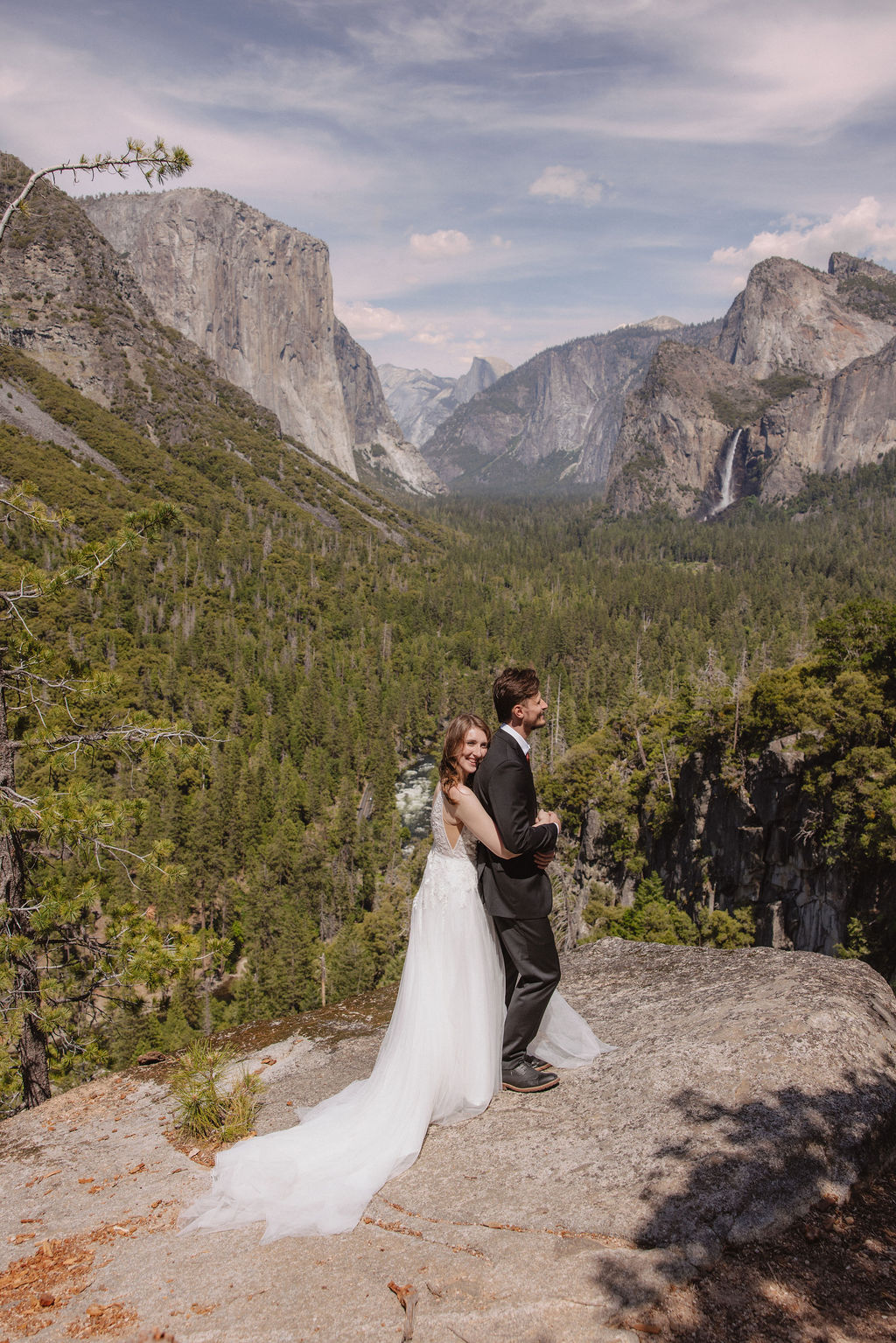 A bride and groom in wedding attire stand on a rocky overlook, embracing while facing a scenic view of mountains, cliffs, and forest in Yosemite National Park.