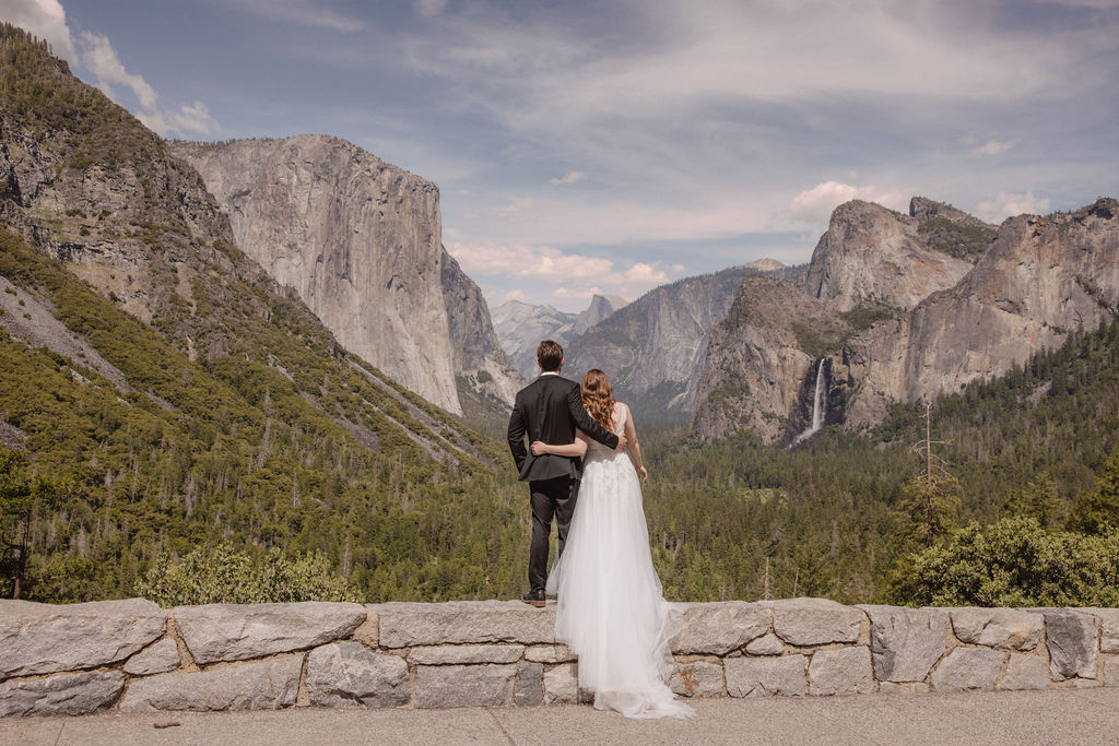 A bride and groom in wedding attire stand on a rocky overlook, embracing while facing a scenic view of mountains, cliffs, and forest in Yosemite National Park.