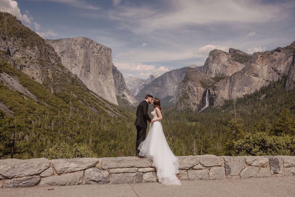 A bride and groom in wedding attire stand on a rocky overlook, embracing while facing a scenic view of mountains, cliffs, and forest in Yosemite National Park.
