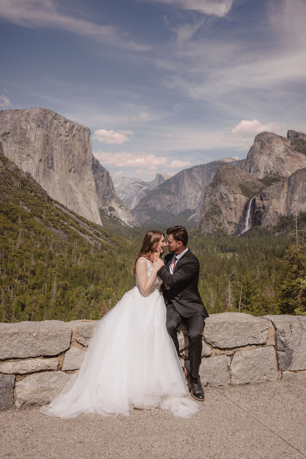 A bride and groom in wedding attire stand on a rocky overlook, embracing while facing a scenic view of mountains, cliffs, and forest in Yosemite National Park.