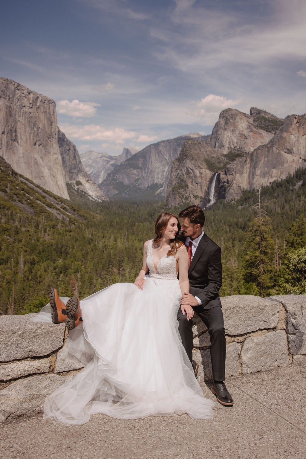 A bride and groom in wedding attire stand on a rocky overlook, embracing while facing a scenic view of mountains, cliffs, and forest in Yosemite National Park.