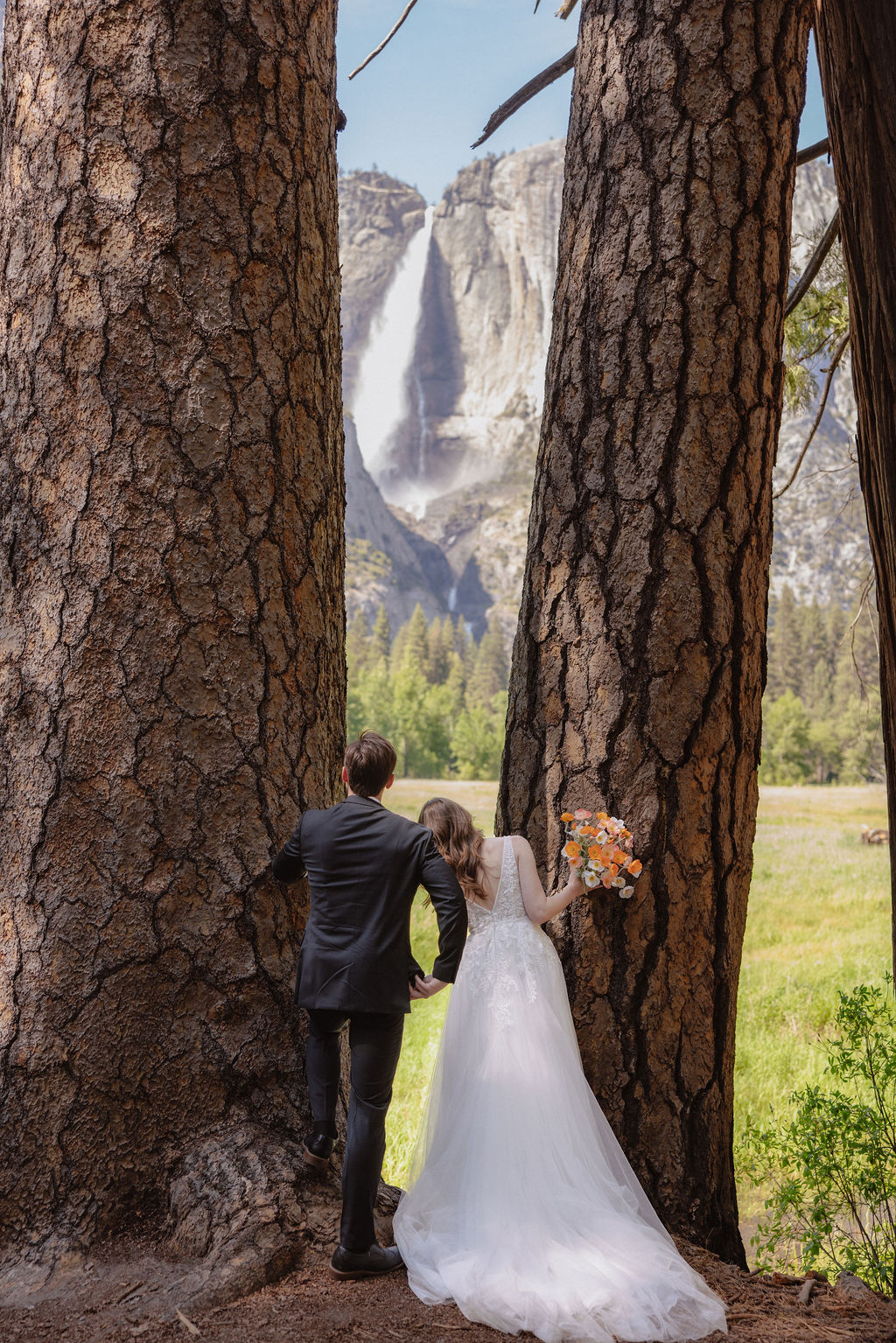 A bride and groom stand hand in hand, facing a green meadow with a tall waterfall and rocky cliffs in the background for a last minute elopement in Yosemite