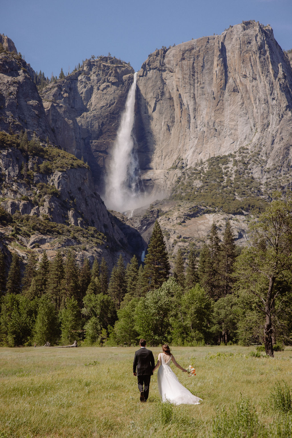 A bride and groom hold hands while walking in a grassy field, with mountains, trees, and a waterfall visible in the background under a partly cloudy sky.