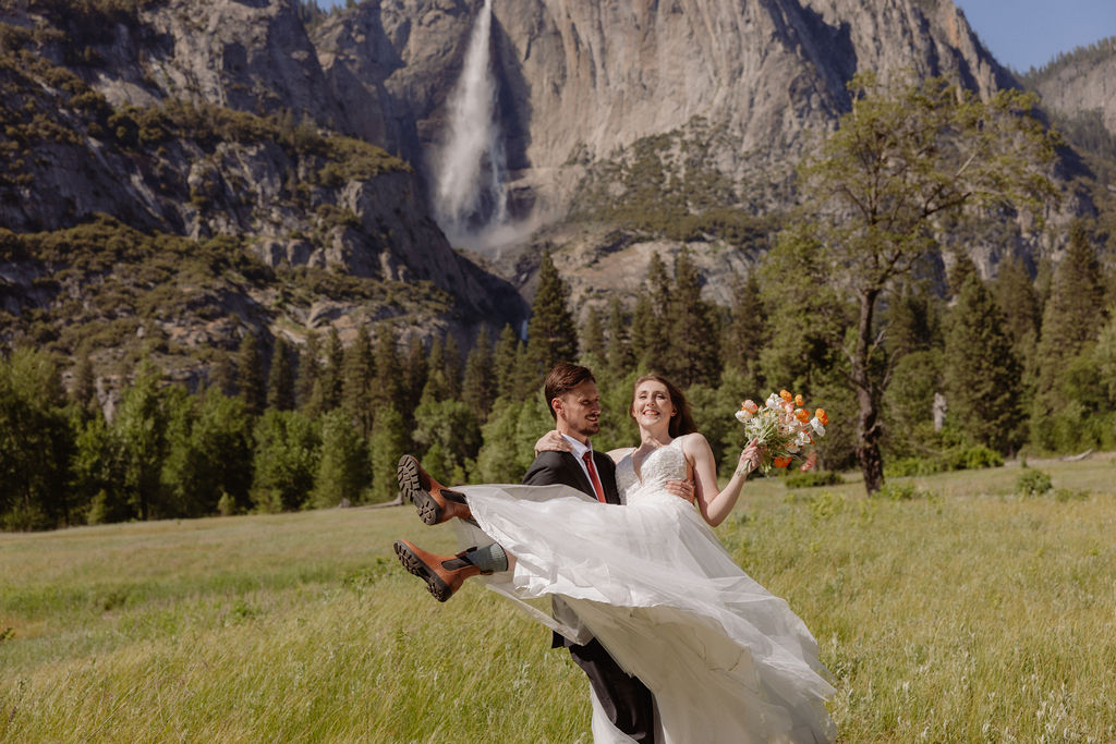 A groom lifts a bride in a grassy field with mountains and a waterfall in the background. The bride holds a bouquet and they both smile.