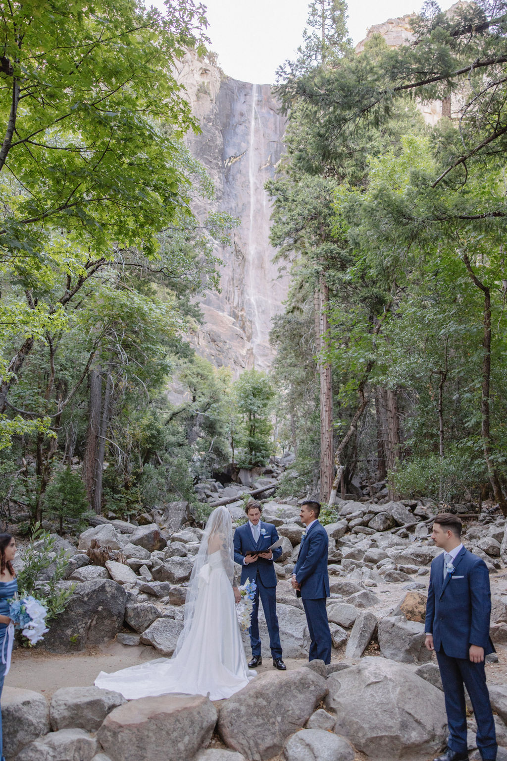 A bride and groom stand with an officiant and two attendants on rocks in a forested area with a waterfall in the background for an adventure elopement