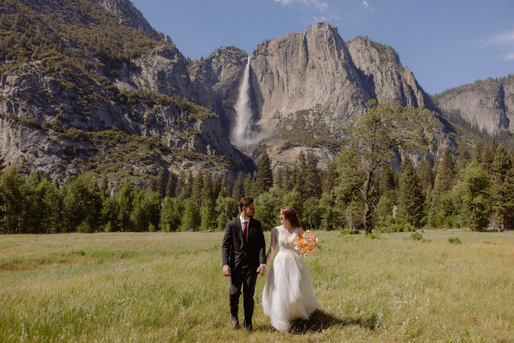 A groom lifts a bride in a grassy field with mountains and a waterfall in the background. The bride holds a bouquet and they both smile.