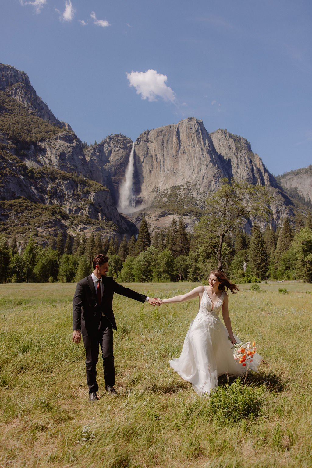 A bride and groom hold hands while walking in a grassy field, with mountains, trees, and a waterfall visible in the background under a partly cloudy sky.