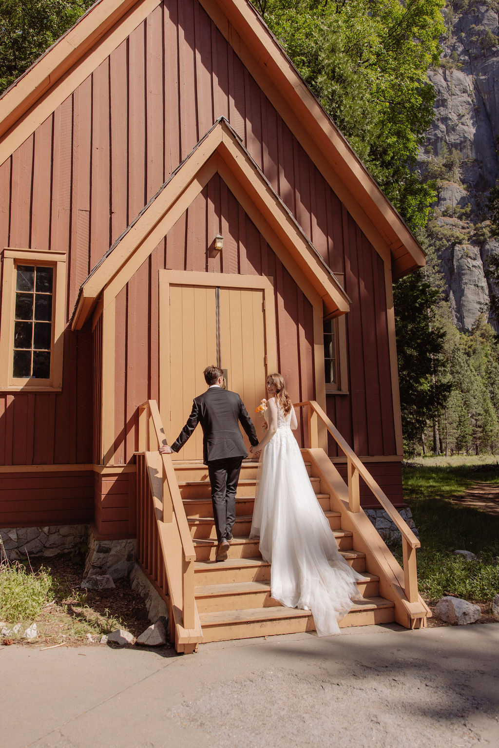 A bride and groom walk up the steps of a small wooden chapel surrounded by trees and sunlight.