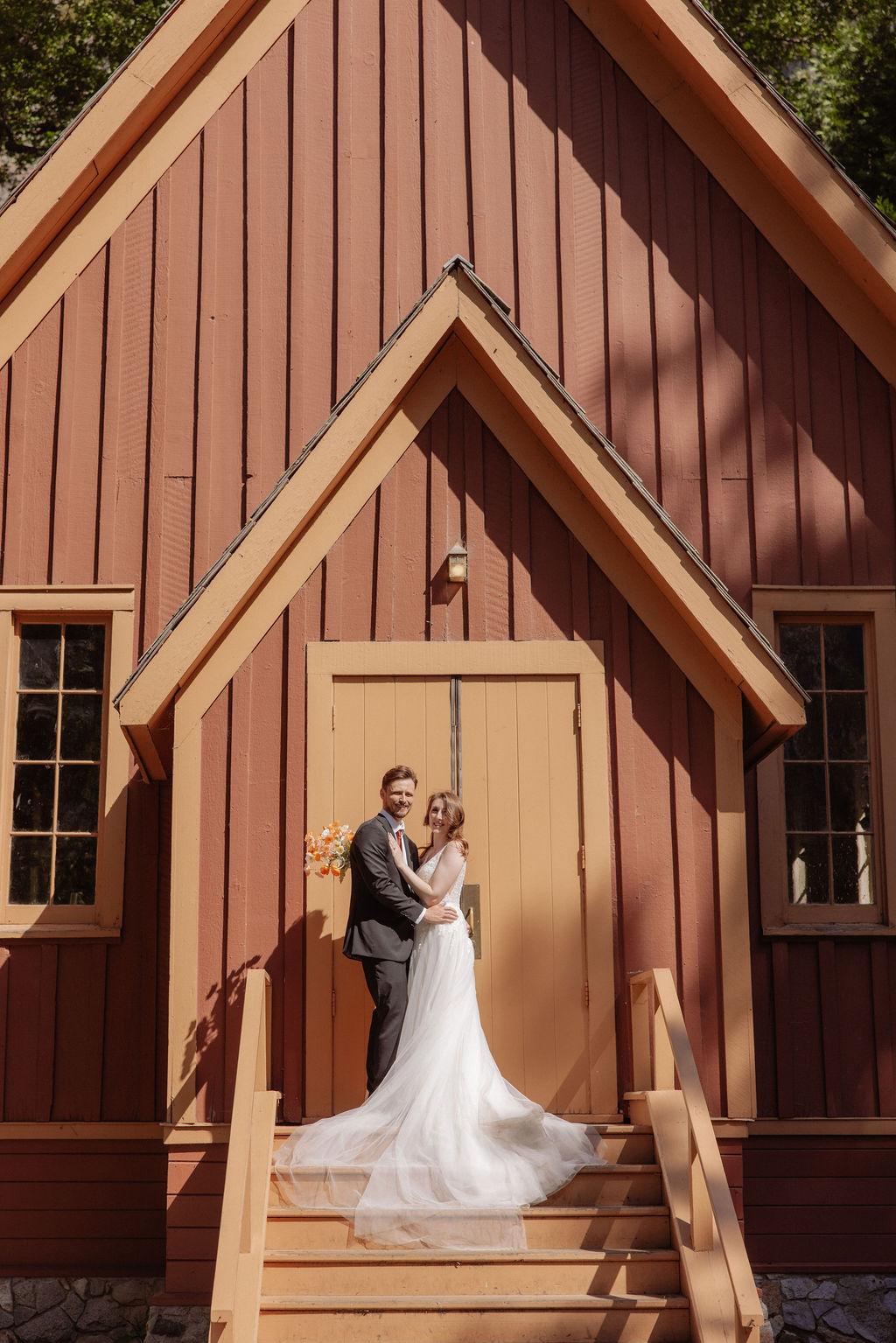 A bride and groom walk up the steps of a small wooden chapel surrounded by trees and sunlight.