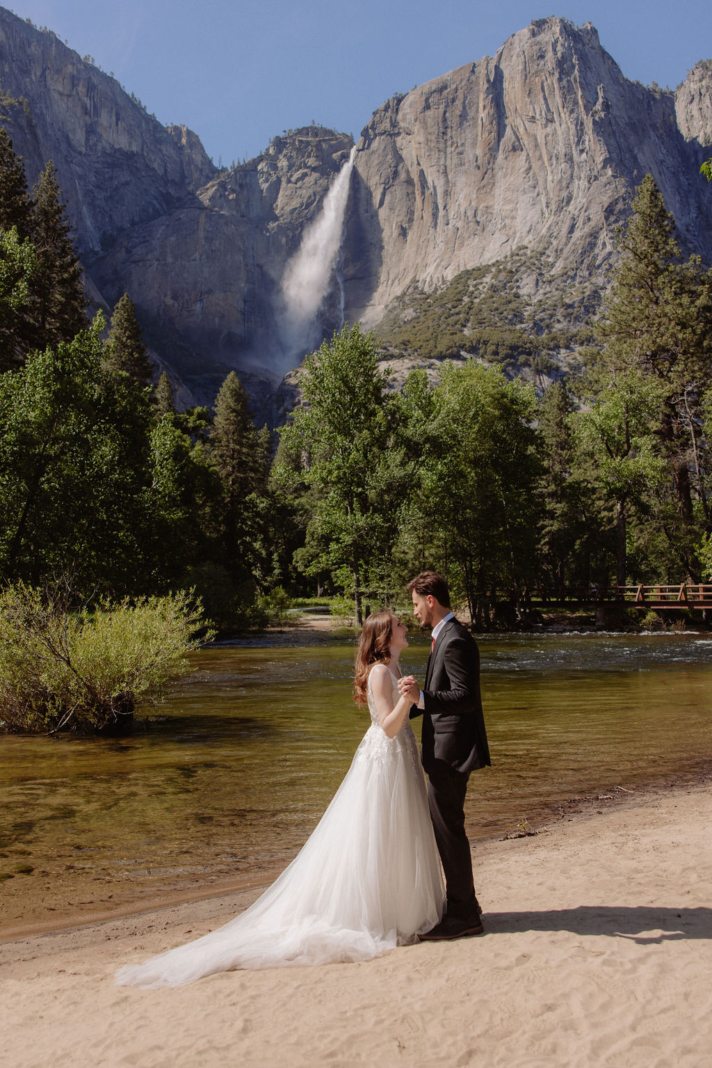 A bride and groom stand together on a sandy riverbank with a waterfall and forested mountains in the background.