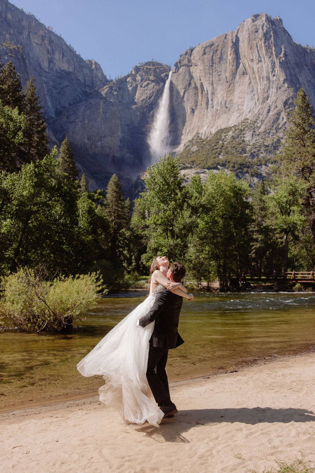 A bride and groom stand together on a sandy riverbank with a waterfall and forested mountains in the background.