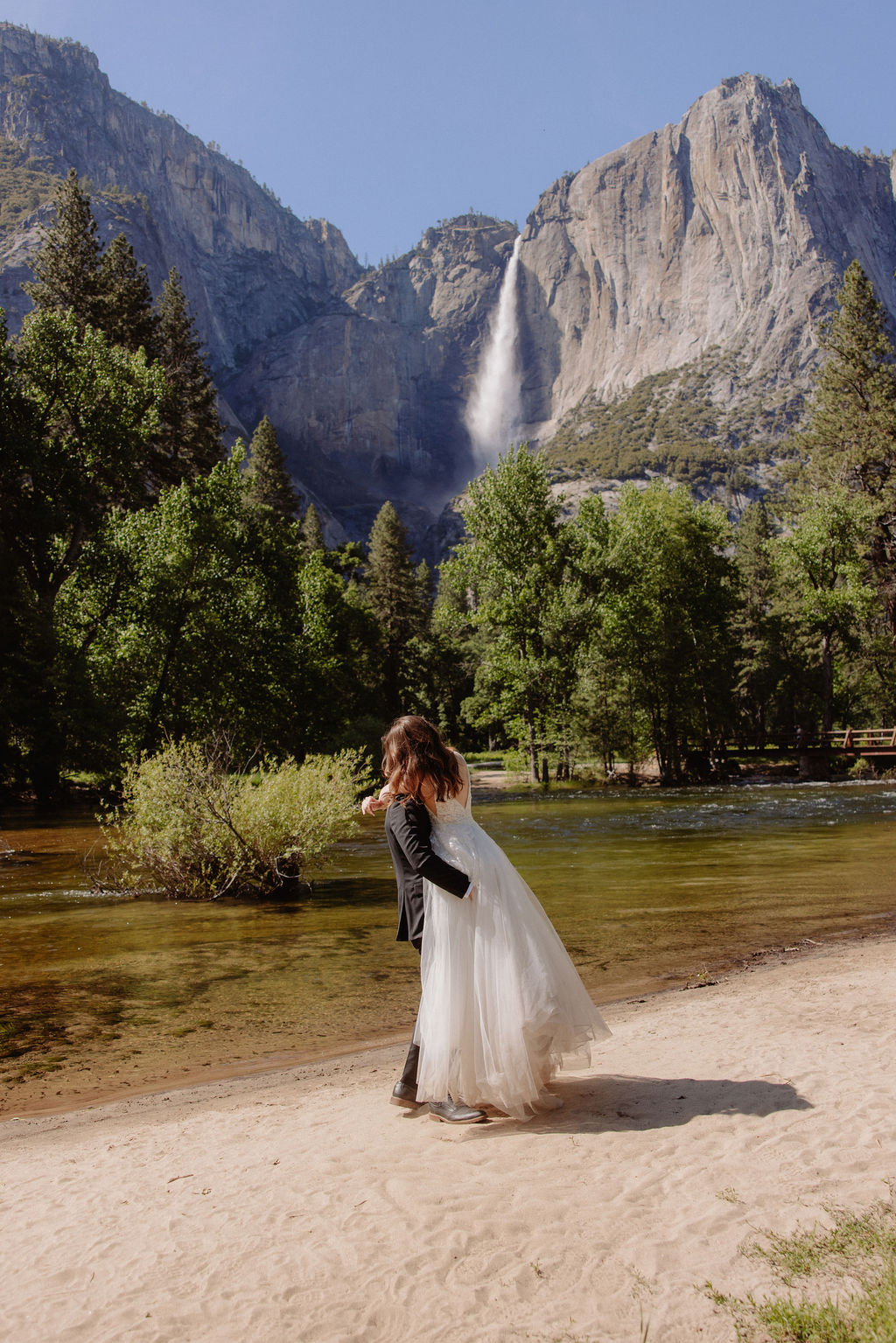 A bride and groom stand together on a sandy riverbank with a waterfall and forested mountains in the background.