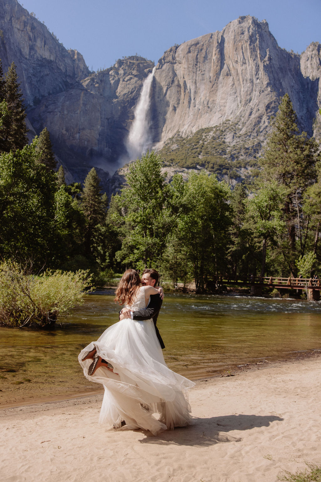 A bride and groom stand together on a sandy riverbank with a waterfall and forested mountains in the background.