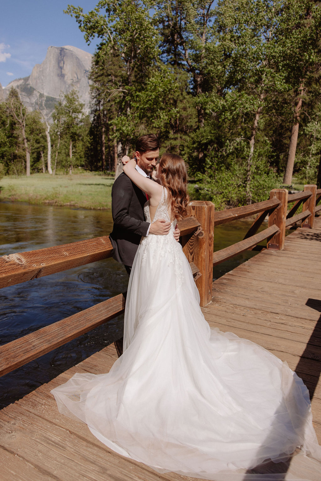 A bride and groom stand together on a sandy riverbank with a waterfall and forested mountains in the background.