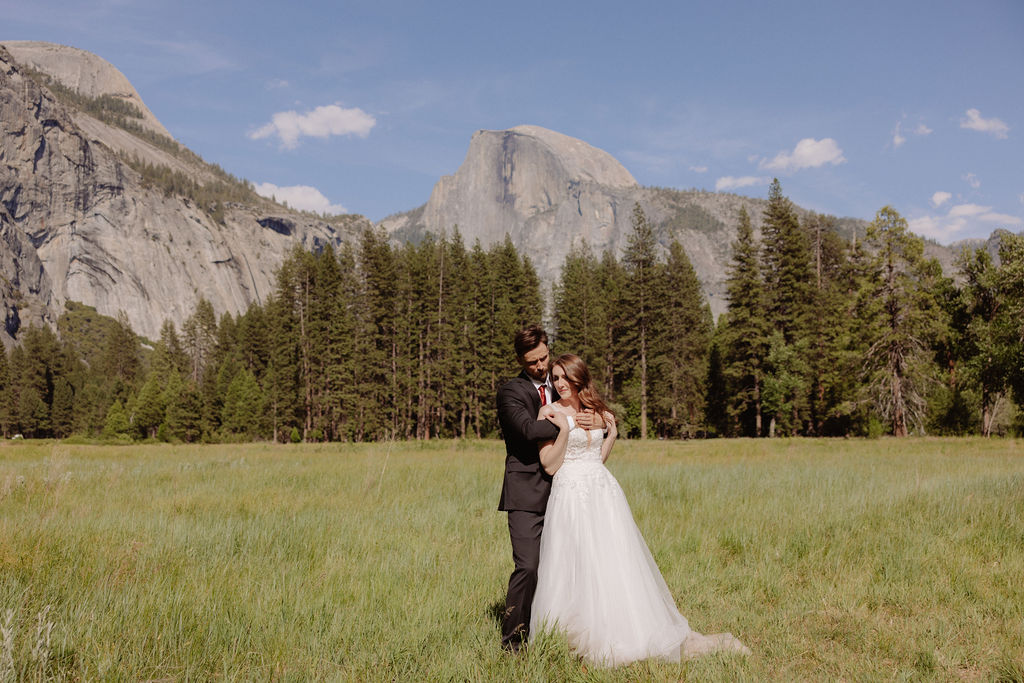 A bride and groom stand together in a grassy field with mountains and pine trees in the background under a clear blue sky for a last minute elopement in Yosemite