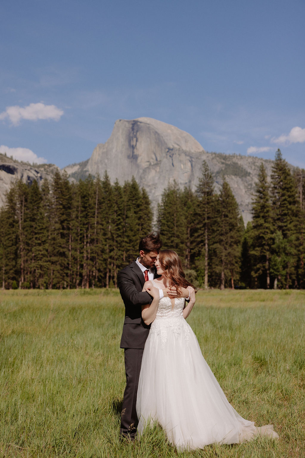 A bride and groom stand together in a grassy field with mountains and pine trees in the background under a clear blue sky for a last minute elopement in Yosemite