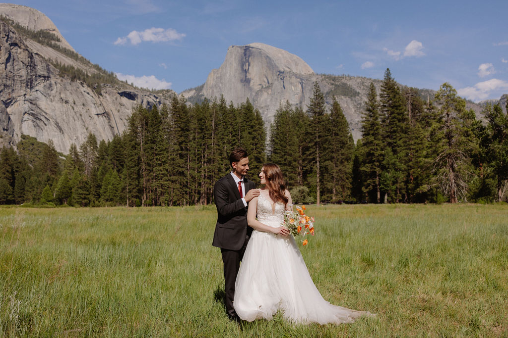 A bride and groom stand together in a grassy field with mountains and pine trees in the background under a clear blue sky for a last minute elopement in Yosemite