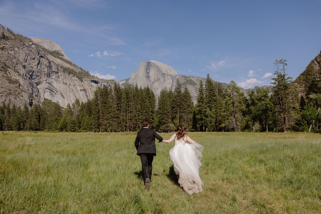 A bride and groom stand together in a grassy field with mountains and pine trees in the background under a clear blue sky for a last minute elopement in Yosemite