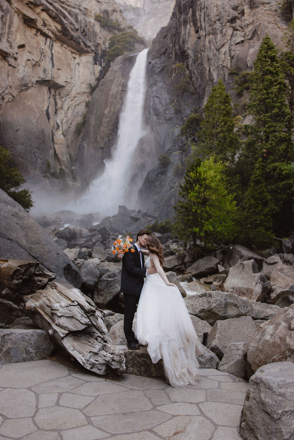A bride and groom stand on rocky terrain in front of a waterfall and tall cliffs, surrounded by trees.
