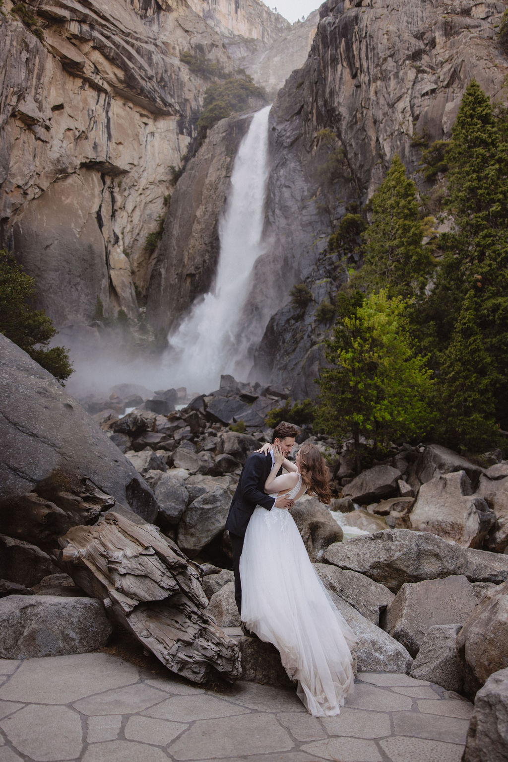 A bride and groom stand on rocky terrain in front of a waterfall and tall cliffs, surrounded by trees.