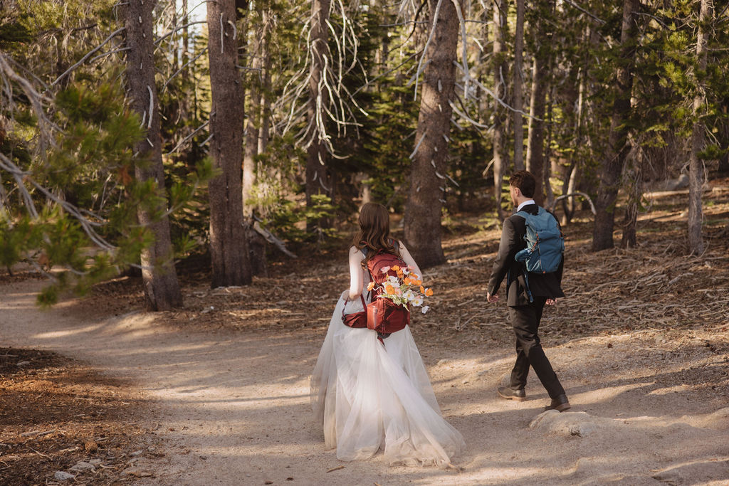 A couple wearing wedding attire and backpacks walk on a dirt trail through a forest, with trees and sunlight in the background.