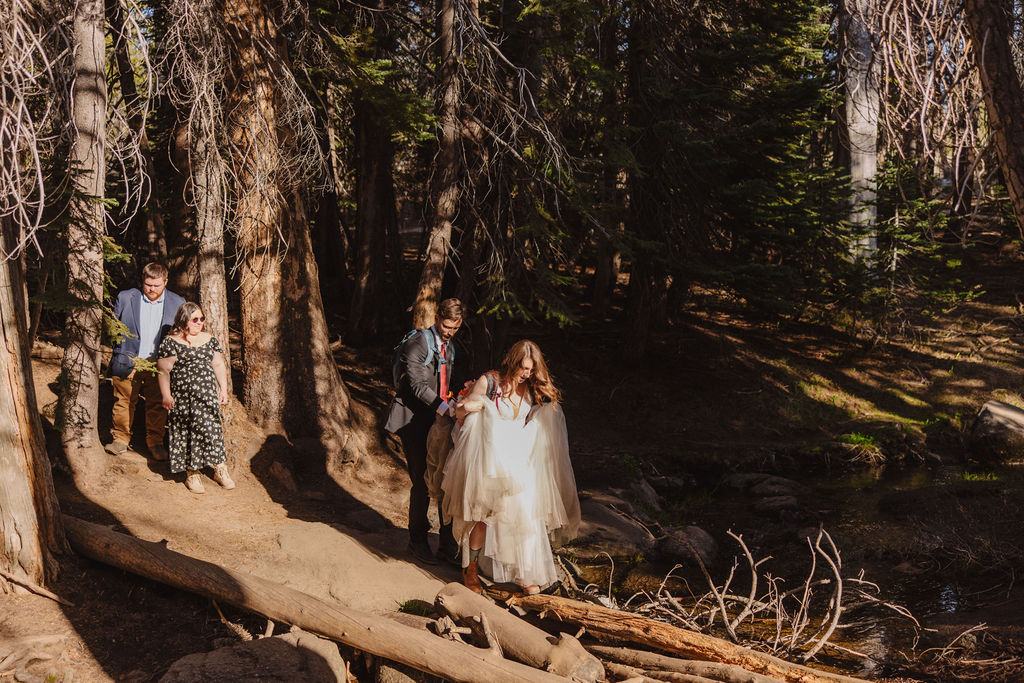A couple wearing wedding attire and backpacks walk on a dirt trail through a forest, with trees and sunlight in the background.