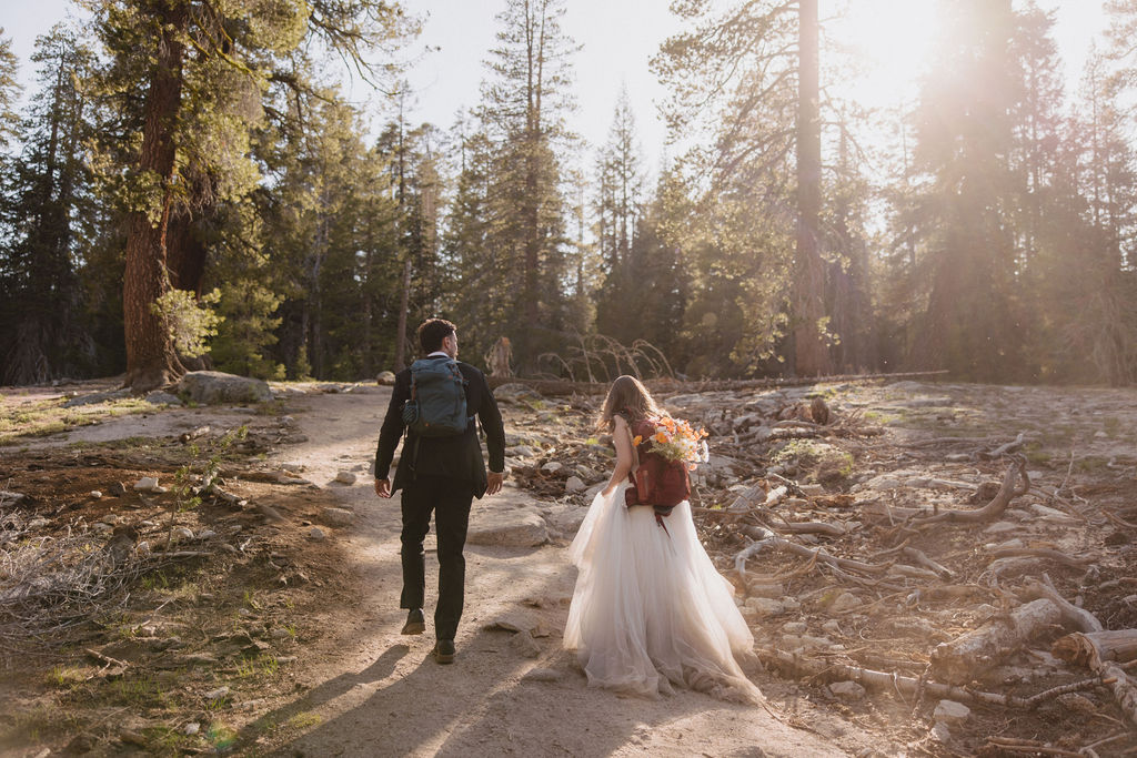 A couple wearing wedding attire and backpacks walk on a dirt trail through a forest, with trees and sunlight in the background.