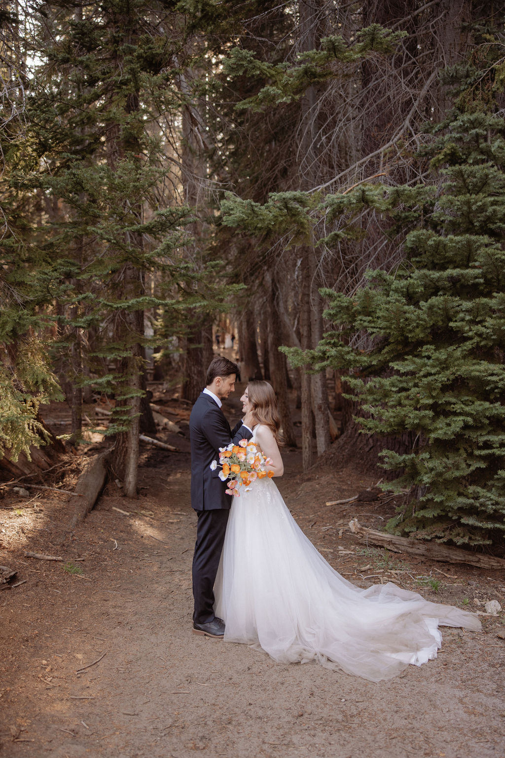 A couple dressed formally stands in a forest, facing each other closely. The woman holds a bouquet of orange and white flowers.