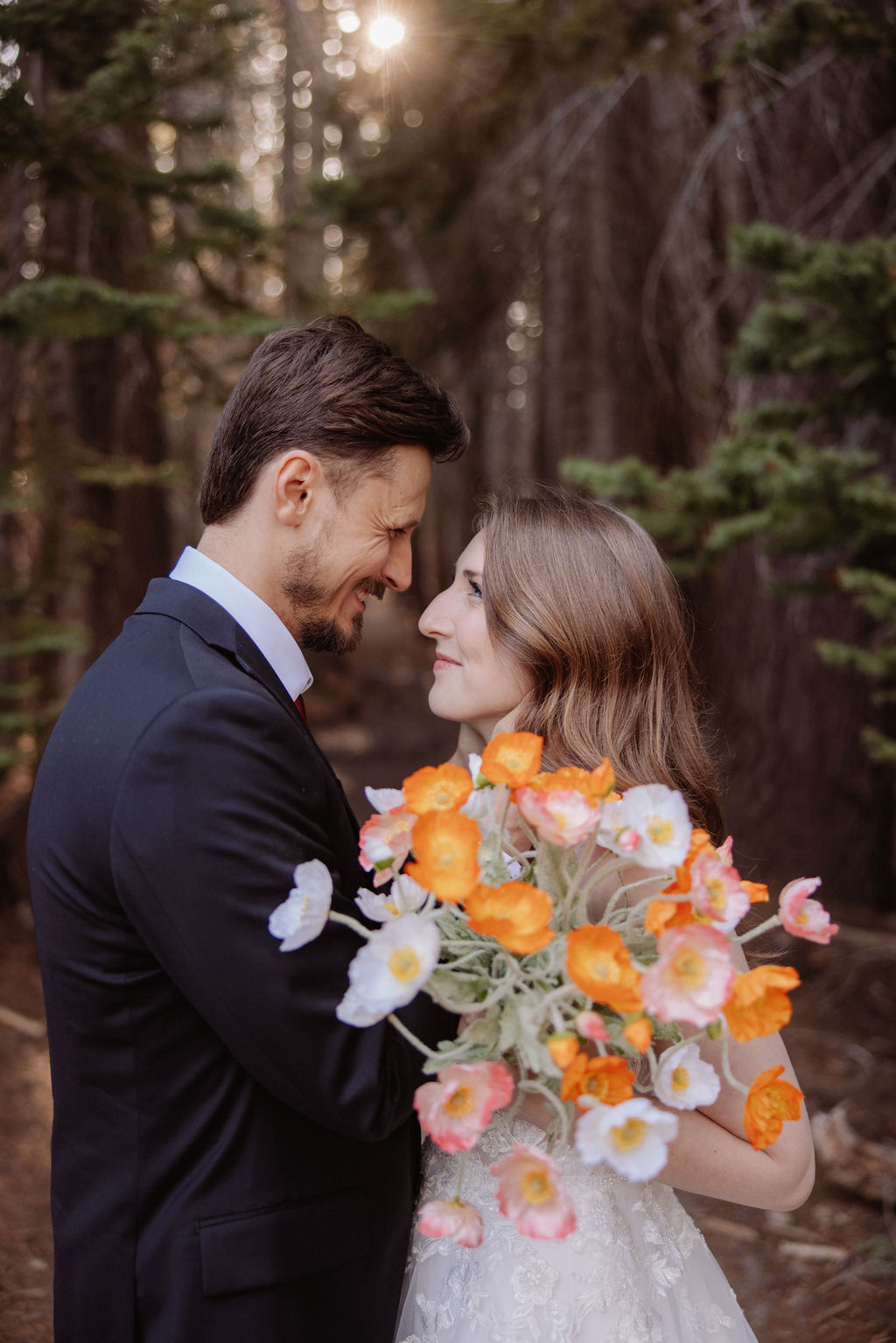 A couple dressed formally stands in a forest, facing each other closely. The woman holds a bouquet of orange and white flowers.