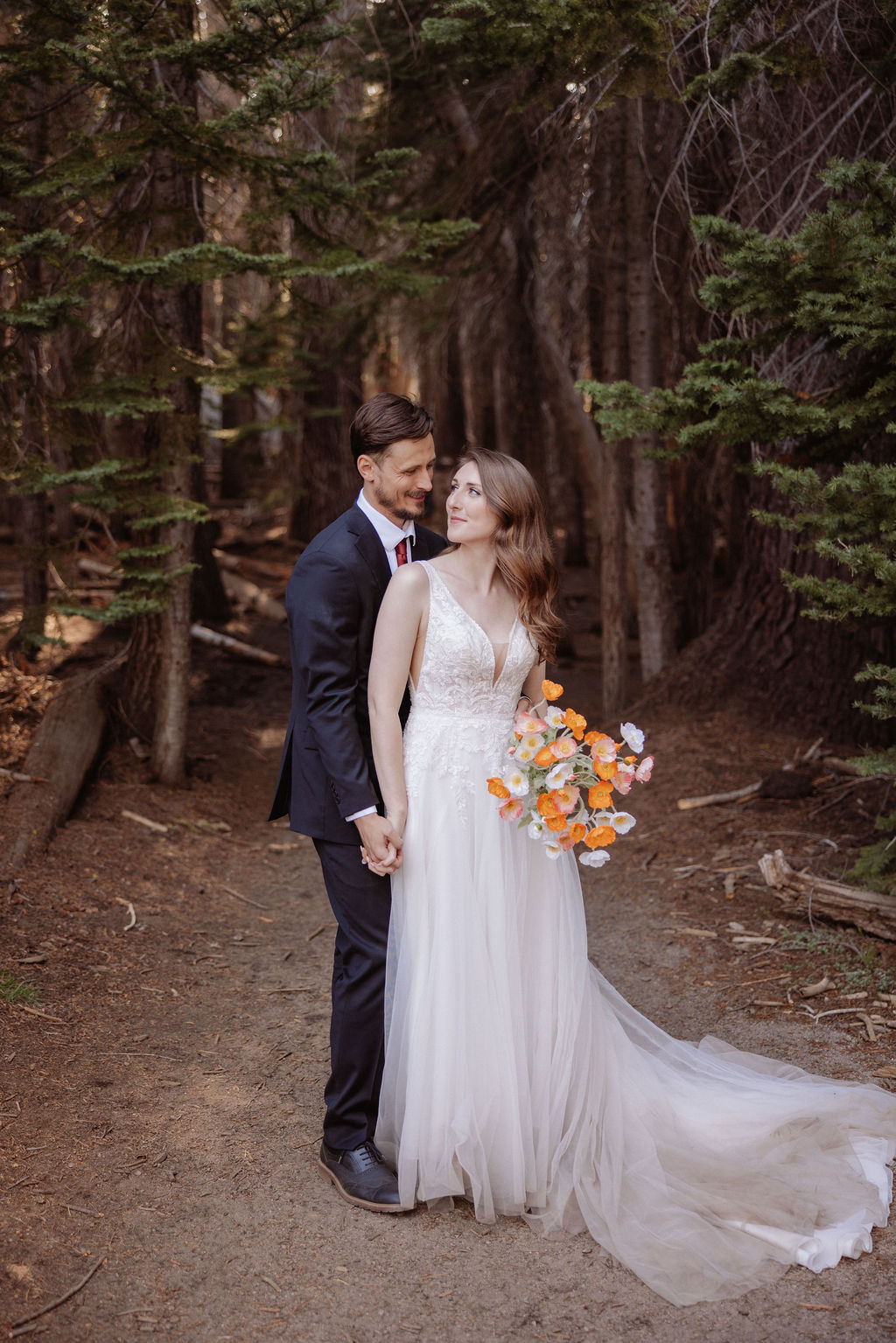 A couple dressed formally stands in a forest, facing each other closely. The woman holds a bouquet of orange and white flowers.