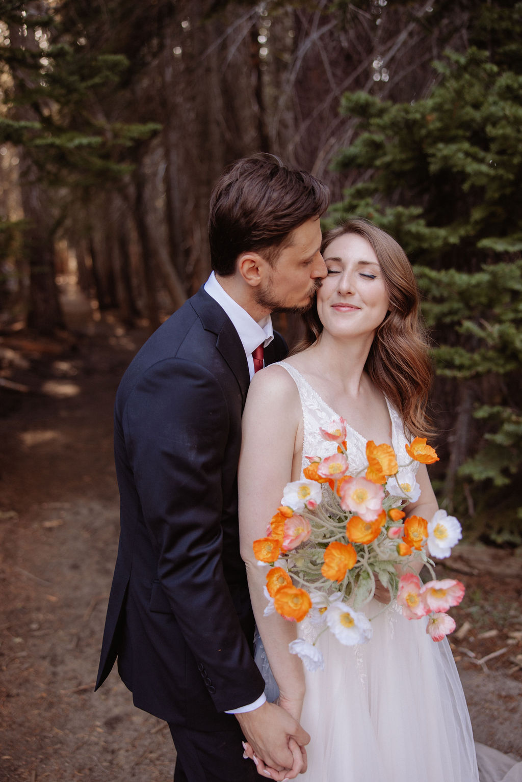 A couple dressed formally stands in a forest, facing each other closely. The woman holds a bouquet of orange and white flowers.