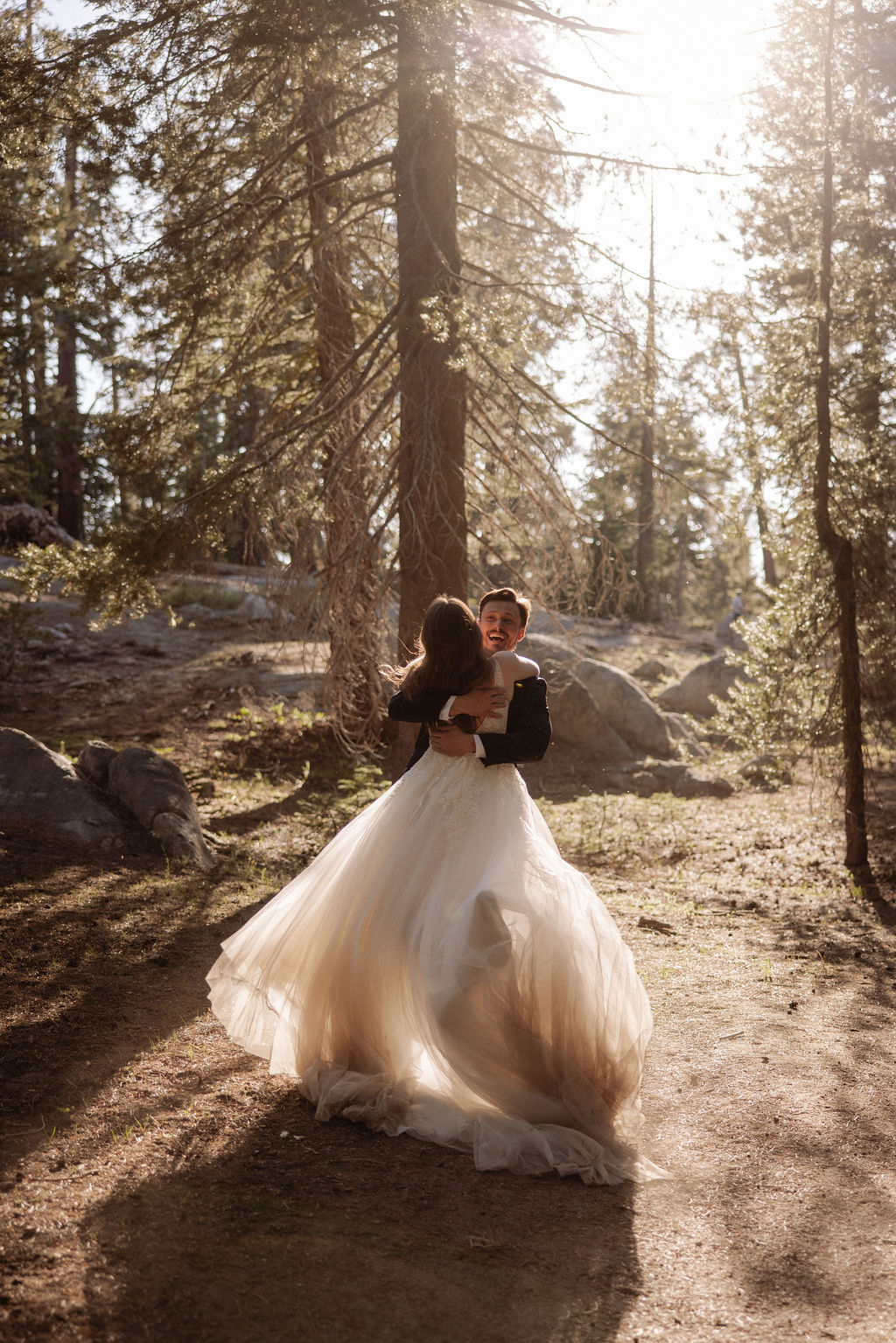 A bride and groom dance together in a sunlit forest, surrounded by tall trees and scattered rocks.