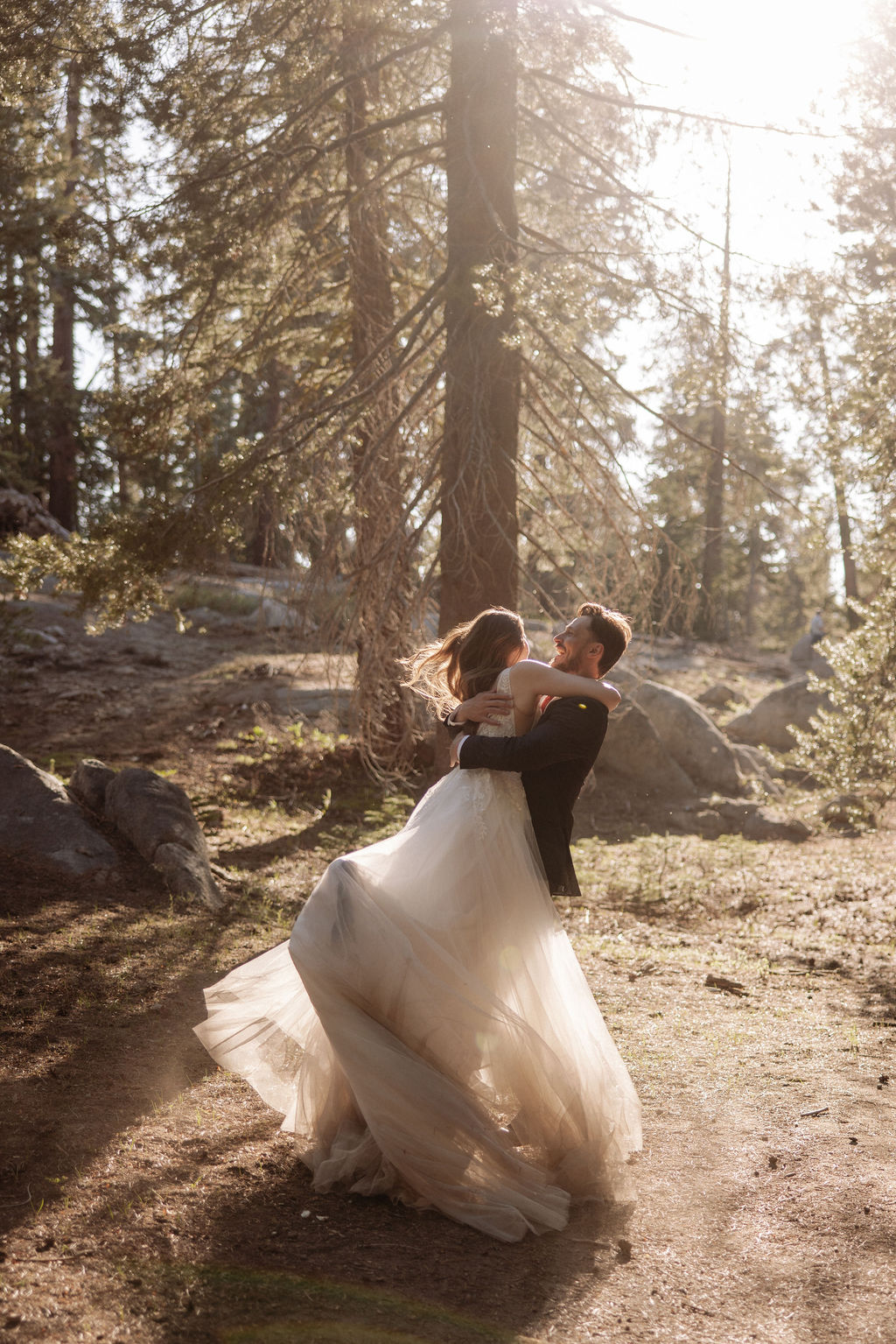 A bride and groom dance together in a sunlit forest, surrounded by tall trees and scattered rocks.