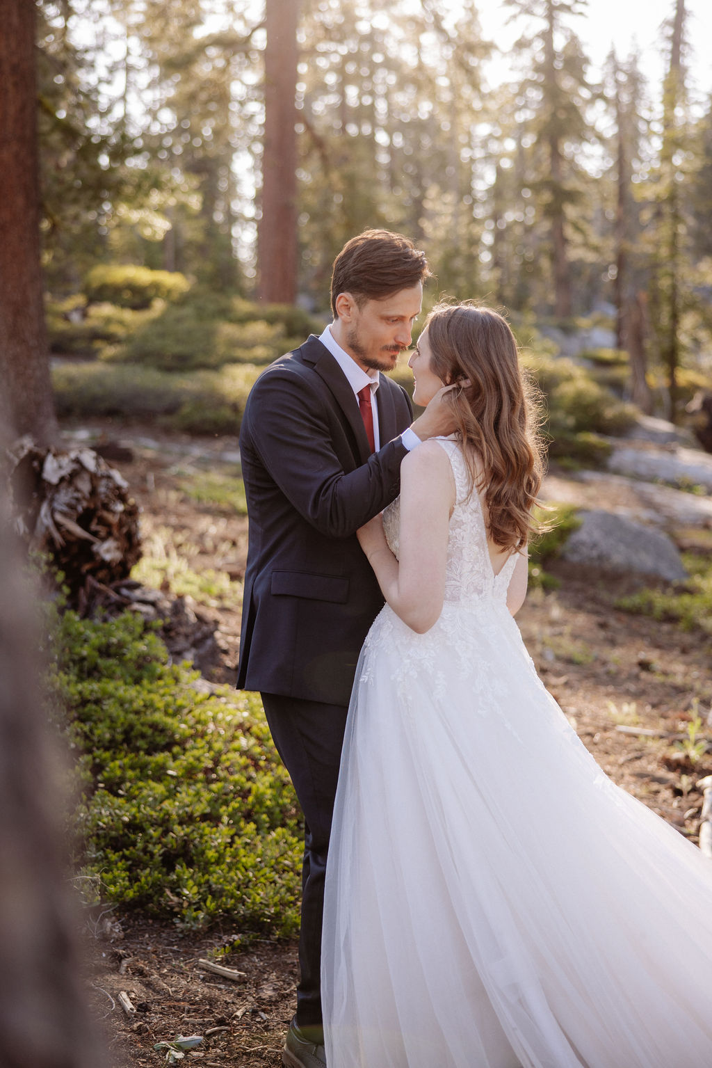 A bride and groom stand close together in a forest, facing each other, with sunlight filtering through the trees.