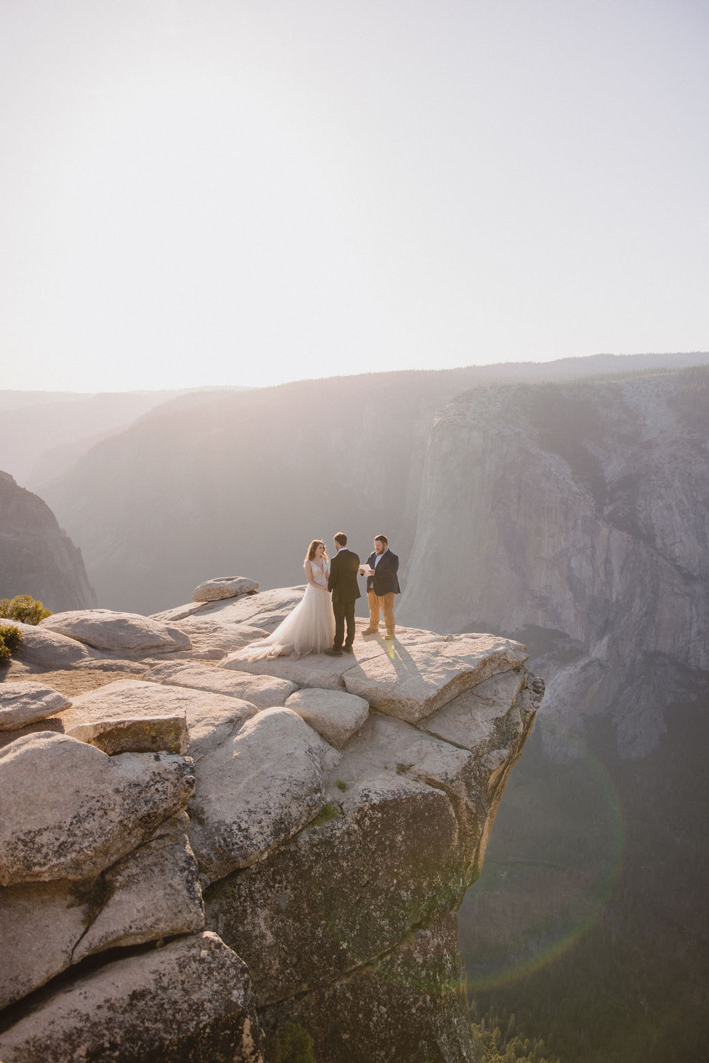 A bride, groom, and officiant stand on the edge of a rocky cliff at sunrise or sunset with mountains in the background.