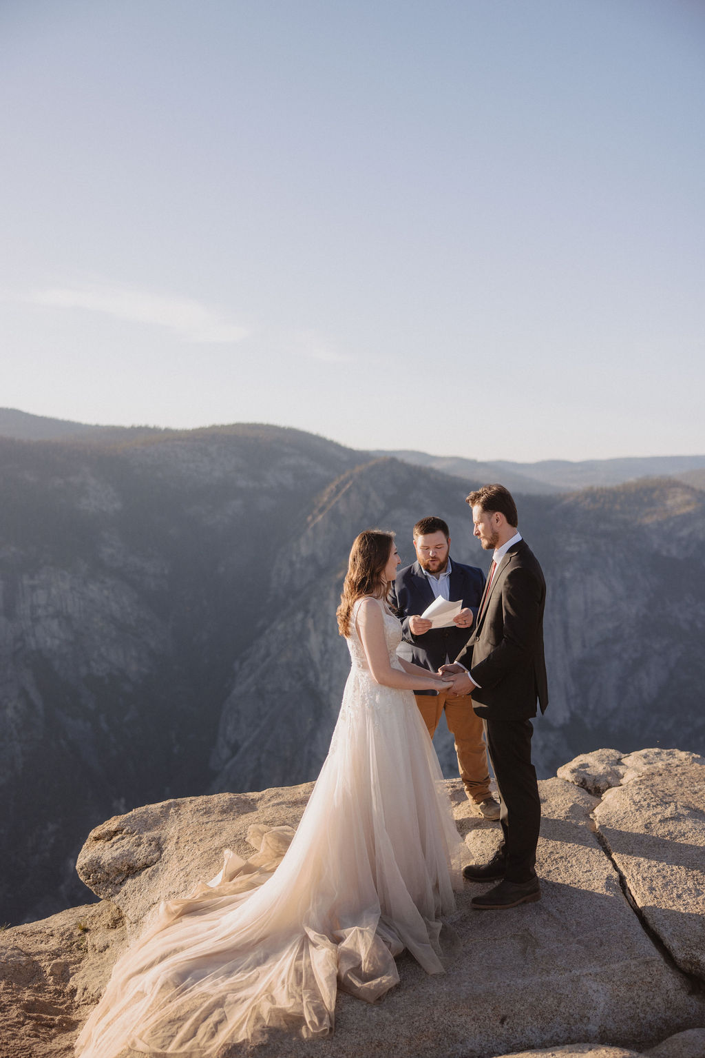 A bride, groom, and officiant stand on the edge of a rocky cliff at sunrise or sunset with mountains in the background.