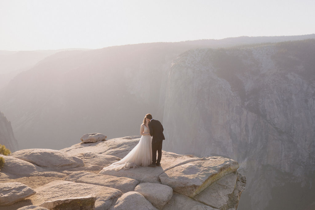 A bride and groom stand together on a rocky cliff edge overlooking a vast, hazy canyon landscape for their last minute yosemite elopement
