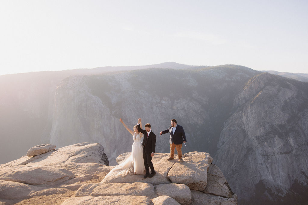 A bride, groom, and officiant stand on the edge of a rocky cliff at sunrise or sunset with mountains in the background.