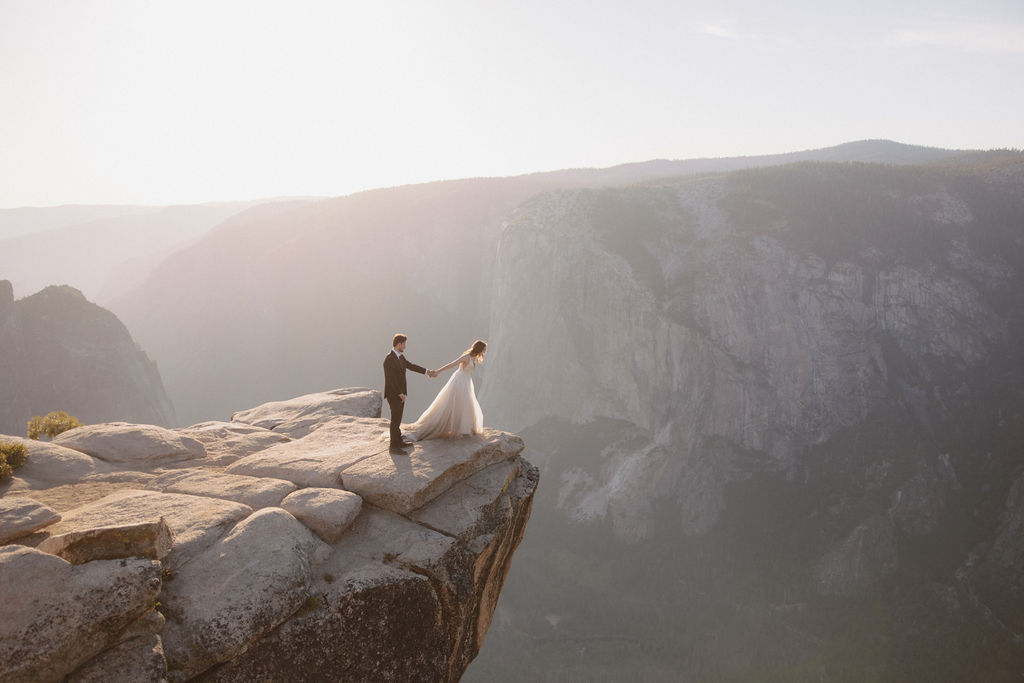 A bride and groom stand hand in hand on the edge of a rocky cliff, overlooking a vast mountainous landscape under a hazy sky for a last minute elopement in Yosemite