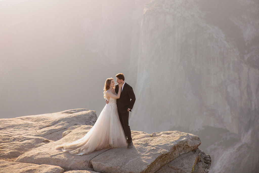 A bride and groom stand together on a rocky cliff edge overlooking a vast, hazy canyon landscape for their last minute yosemite elopement