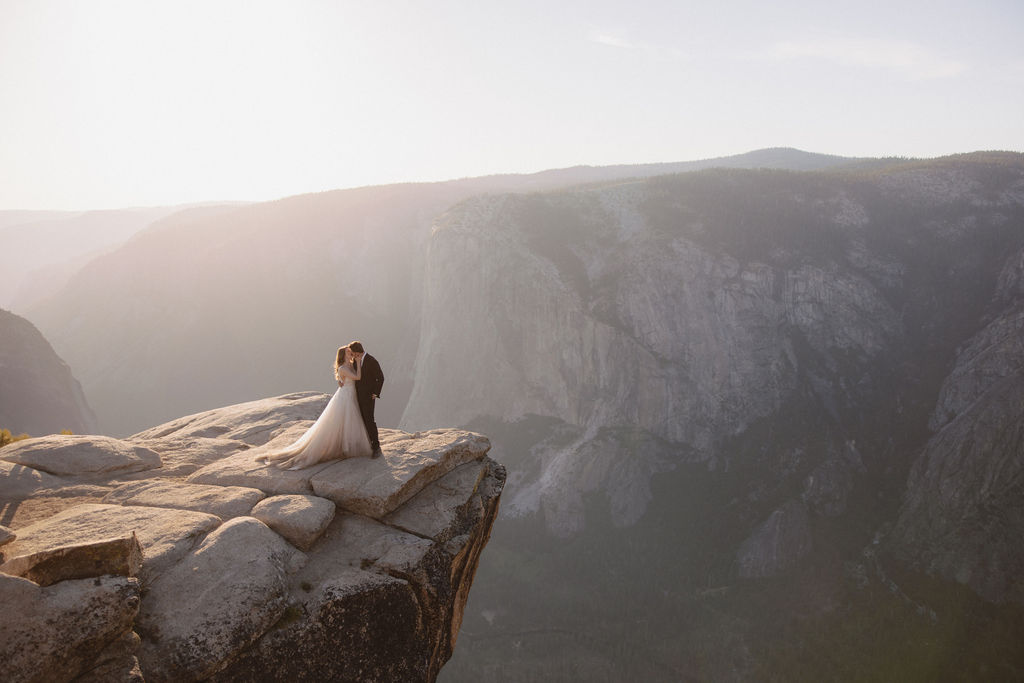 A bride and groom stand hand in hand on the edge of a rocky cliff, overlooking a vast mountainous landscape under a hazy sky for a last minute elopement in Yosemite