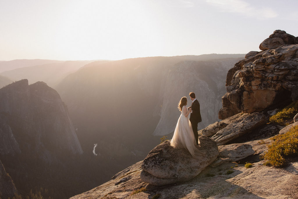 A couple in wedding attire stands on a rocky cliff at sunset, overlooking a vast mountainous landscape for a last minute elopement in Yosemite
