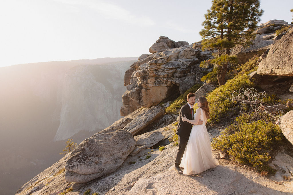 A couple in wedding attire stands on a rocky cliff at sunset, overlooking a vast mountainous landscape for a last minute elopement in Yosemite