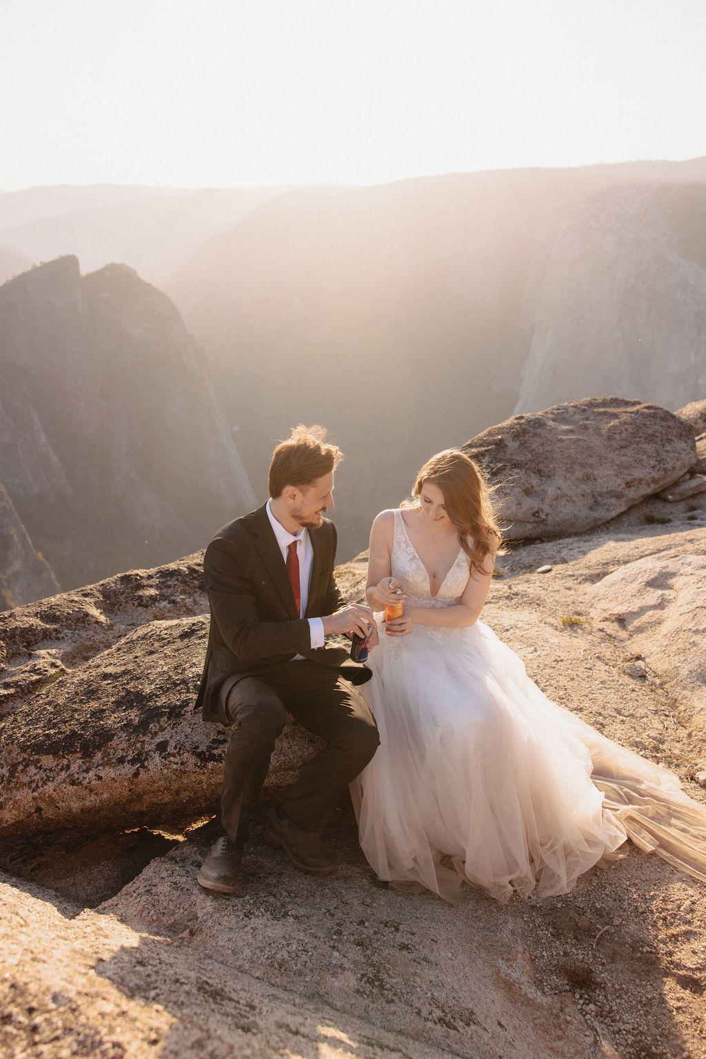 A couple dressed in wedding attire sits on a rocky cliff at sunset, holding glasses and looking at each other with mountains in the background for a last minute elopement in Yosemite