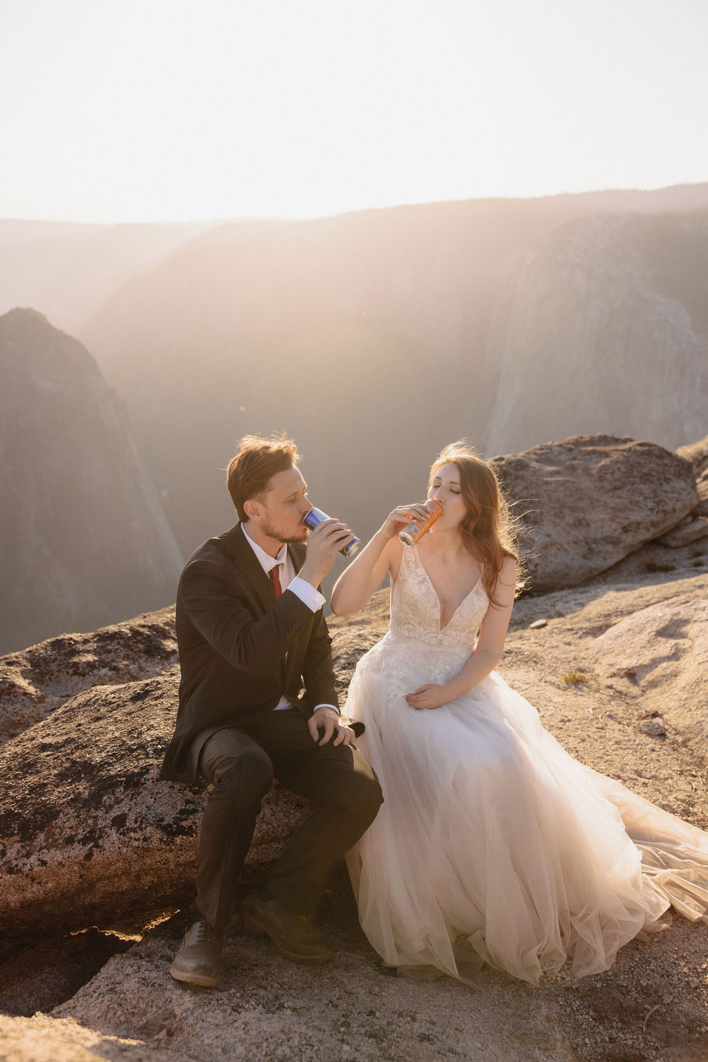 A couple dressed in wedding attire sits on a rocky cliff at sunset, holding glasses and looking at each other with mountains in the background for a last minute elopement in Yosemite