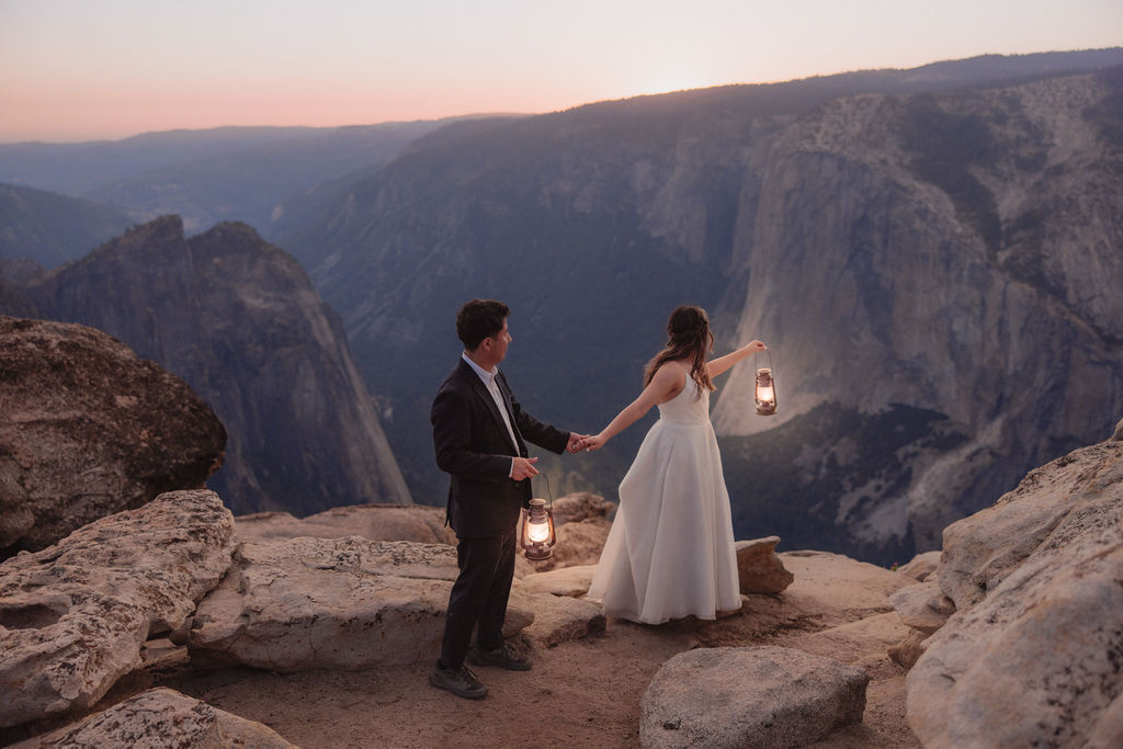 A couple in formal attire holds lanterns while standing on rocky cliffs at sunset, overlooking a vast mountain landscape.