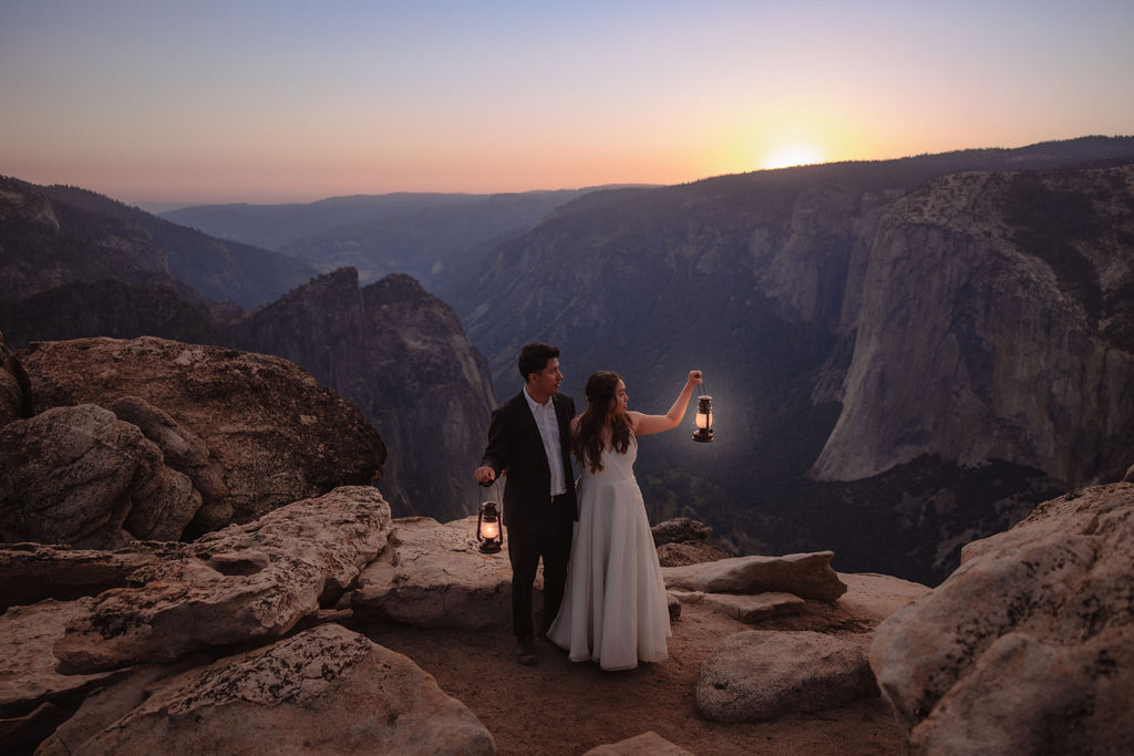 A couple in formal attire holds lanterns while standing on rocky cliffs at sunset, overlooking a vast mountain landscape.