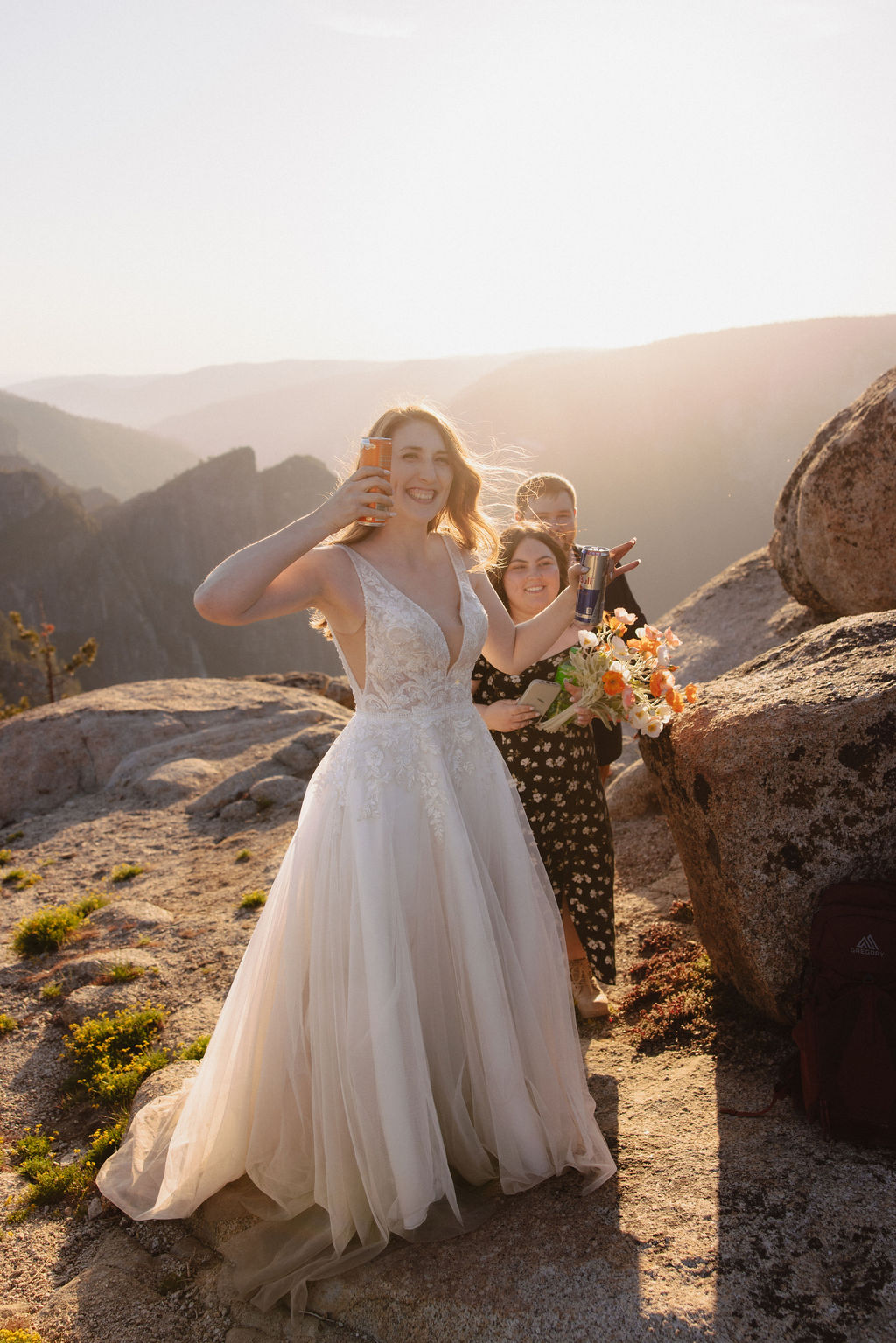 A couple dressed in wedding attire sits on a rocky cliff at sunset, holding glasses and looking at each other with mountains in the background for a last minute elopement in Yosemite